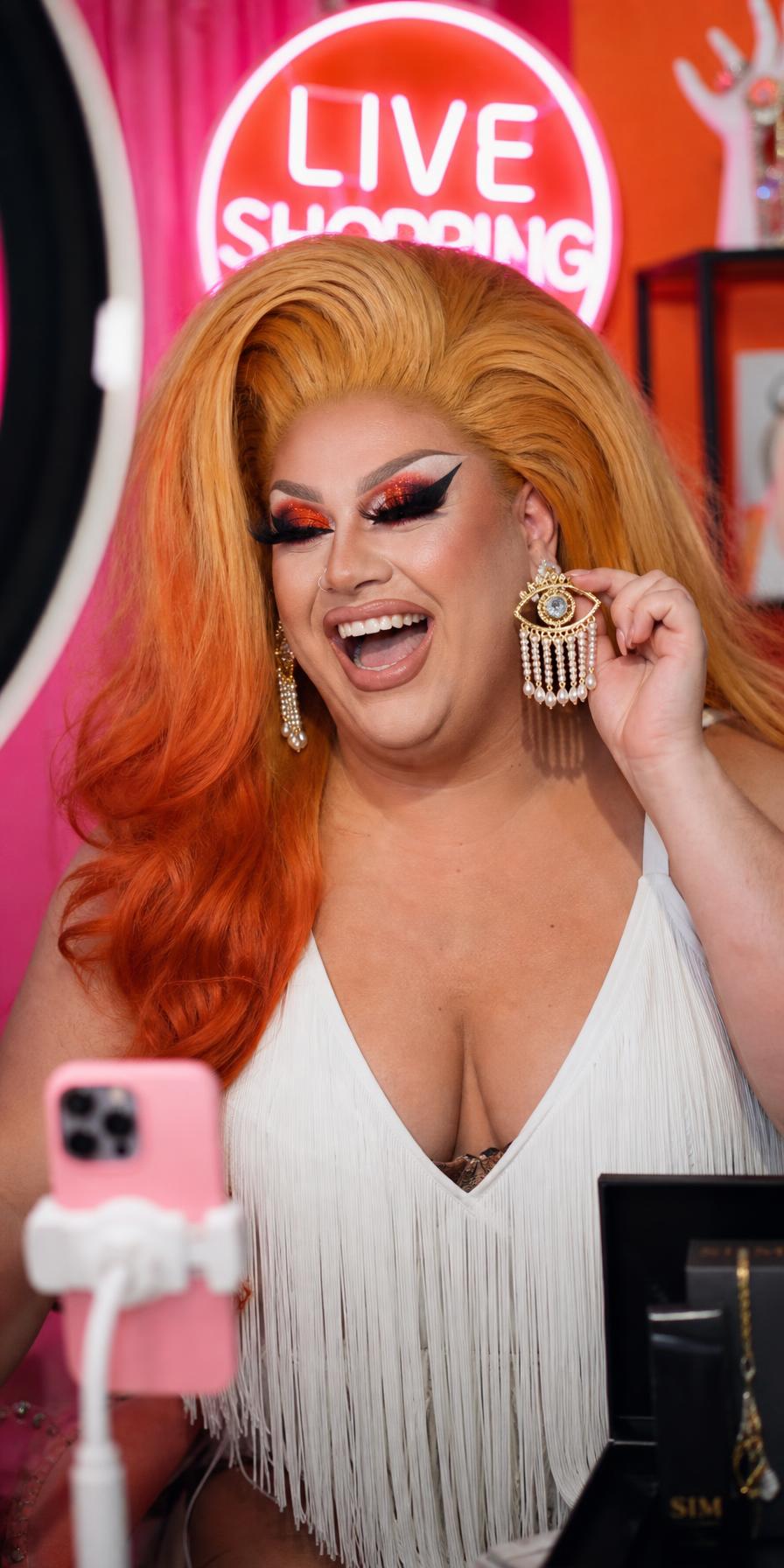 A drag queen host laughing mid-broadcast while holding up a statement earring